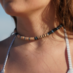 Bluefin heishi choker of beige, sky blue, and black heishi beads on neck of model with blue and white swimsuit straps and blue sky behind her.