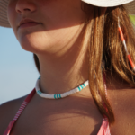 Surf's Up necklace designed with white & turquoise heishi beads in traditional surfer pattern on model model in hat.