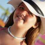 Surf's Up necklace designed with white & turquoise heishi beads in traditional surfer pattern on smiling model in hat.