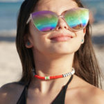 Volcanic Necklace of white, black, red, yellow, & turquoise heishi beads on model in sunglasses with sand & water in the background.