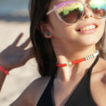 Volcanic Necklace of white, black, red, yellow, & turquoise heishi beads on the neck of a smiling model in sunglasses with sand & water in the background.