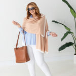 Dreamsoft Organic Cotton Travel Scarf Blush pink in color with white band shown as wrap on smiling model in white pants & light blue shirt holding a brown purse & wearing sunglasses in front of white decorative wall with green plant on the side.