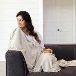 Dreamsoft Organic Cotton Travel Scarf in neutral color used as blanket on dark haired smiling model sitting on dark colored couch holding small wooden bowl