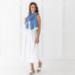 Dreamsoft Organic Cotton Travel Scarf French Blue in color with white band shown as scarf on smiling model in white dress & sandals in front of white decorative wall.