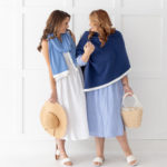 Dreamsoft Organic Cotton Travel Scarf French Blue in color with white band shown as scarf on model in white dress & sandals holding sun hat in front of white decorative wall laughing with another model in Dreamsoft Travel Scarf Navy in color with white band wearing light blue dress & sandals holding a basket bag.