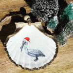 Santa's Sea Creatures Pelican large seashell ornament with painted pelican in Santa hat. Ornament is trimmed in gold leaf and displayed on wooden background with small model display christmas tree accent.