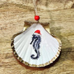 Santa's Sea Creatures Seahorse large seashell ornament with painted seahorse in santa hat. Ornament is trimmed in gold leaf and displayed on wood background.