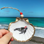 Santa's Sea Creatures Sea Turtle large seashell ornament with painted sea turtle in santa hat. Ornament is trimmed in gold leaf and being held up with beach in the background.