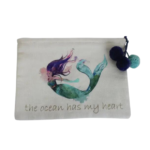 Mermaid Zip Pouch - Ocean Heart displayed on white background. Off white 8" x 6" white cotton zipper bag with screen printed image of mermaid with purple and pink hair and green and blue tail. Zipper is accented with 2 purple and 1 light blue pom poms. Beneath mermaid is message written in gold lowercase letters reading the ocean has my heart.