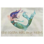 Mermaid Zip Pouch - Ocean Heart close up view of mermaid and writing displayed on white background. Off white 8" x 6" white cotton zipper bag with screen printed image of mermaid with purple and pink hair and green and blue tail. Zipper is accented with 2 purple and 1 light blue pom poms. Beneath mermaid is message written in gold lowercase letters reading the ocean has my heart.