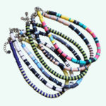 Mythos Collection of heishi necklaces displayed on white background. Multiple necklaces styles with various colors including pink, turquoise, purple, yellow, beige, black, white, and green