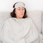 Travel Set Light Gray eye mask and blanket displayed on sleeping model with dark hair lying on light gray couch.