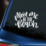 Meet Me At the Beach Vinyl Decal white decal of script lettering displayed on back window of blue car with dark tinting.