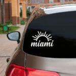 Miami Sun Vinyl Decal white decal with half sun outline with Miami in script beneath displayed on back window of silver hatchback car with dark tinting.