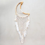 Moon Medium Macrame Dreamcatcher displayed hanging against a light gray backdrop. Hoop on dreamcacher is natural color rattan. Moon and tassels are white cotton macrame. Tassels are finished with white goose feathers.