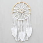 Sunburst Large Macrame Dreamcatcher displayed hanging against a light gray backdrop. Hoop on dreamcacher is natural color rattan. Sunburst and tassels are white cotton and finished with oversized stylized feather made of cotton.